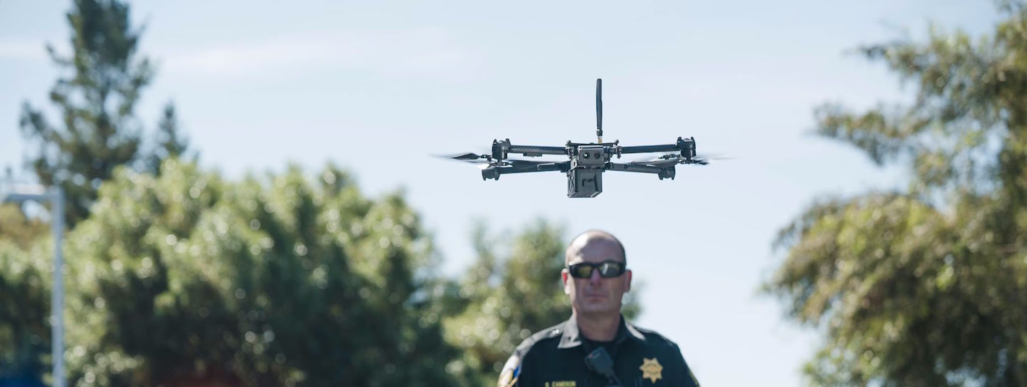 A police drone hovers in the foreground on a sunny day. A police officer stands behind it and next to a vehicle with its emergency lights on.